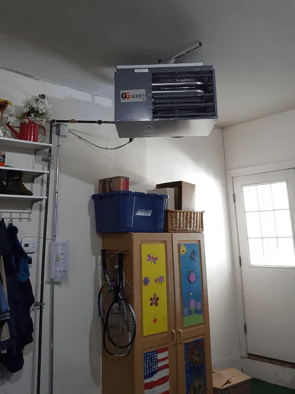 The garage interior features a wall-mounted heater above a cabinet adorned with floral and flag motifs. Tennis rackets hang from the side, while a blue storage bin rests on top, ensuring both style and efficient garage heating in this well-organized space.